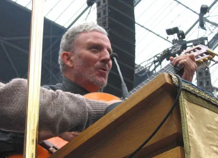 Un uomo dai capelli grigi canta al microfono accompagnandosi con la chitarra, da un palco coperto