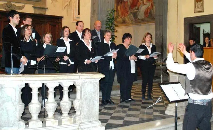 Un coro in abiti neri e camicia bianca canta leggendo gli spartiti nel presbiterio di una chiesa, guidato da un direttore ripreso di spalle