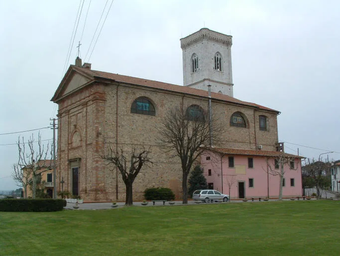 Fianco della chiesa di Orentano con grandi finestre ad arco e il campanile sullo sfondo; a destra un edificio rosa addossato alla chiesa e un prato in primo piano