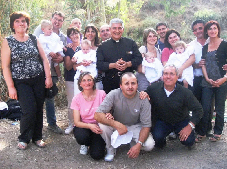 Genitori e familiari con quattro bambini piccoli vestiti di bianco, insieme a un sacerdote, in posa tra i canneti in riva al fiume