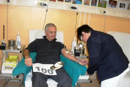 Un donatore con il cartello «100» sulle gambe, seduto sulla poltrona del centro trasfusionale mentre un'operatrice gli prepara il braccio