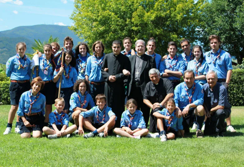Foto di gruppo di scout in uniforme azzurra e fazzolettone con un vescovo con croce pettorale e due sacerdoti, su un prato con colline sullo sfondo