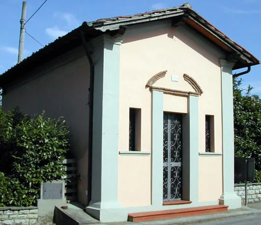 Piccolo oratorio intonacato color rosa chiaro con lesene bianche, tetto a capanna e porta con cancello in ferro battuto, tra le siepi