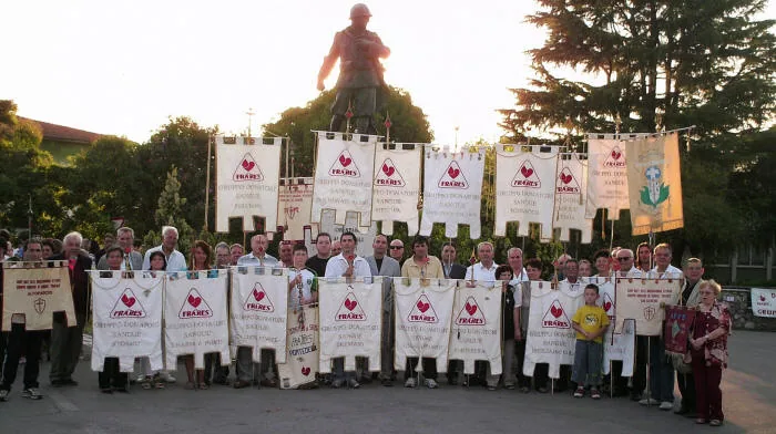 Donatori Fratres in posa all'aperto con numerosi stendardi dei gruppi Fratres, davanti a un monumento sormontato da una statua, al tramonto