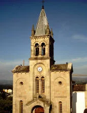 Facciata in pietra di una chiesa con orologio e campanile a cuspide, illuminata dalla luce calda della sera sotto un cielo azzurro