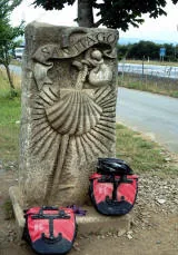 Cippo di pietra con la conchiglia scolpita e la scritta Santiago, con due borse rosse e un casco appoggiati alla base