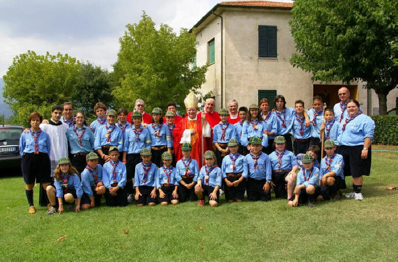 Foto di gruppo di scout in uniforme azzurra e fazzolettone con un vescovo in paramenti rossi con mitra e pastorale e alcuni sacerdoti, sul prato davanti a un edificio